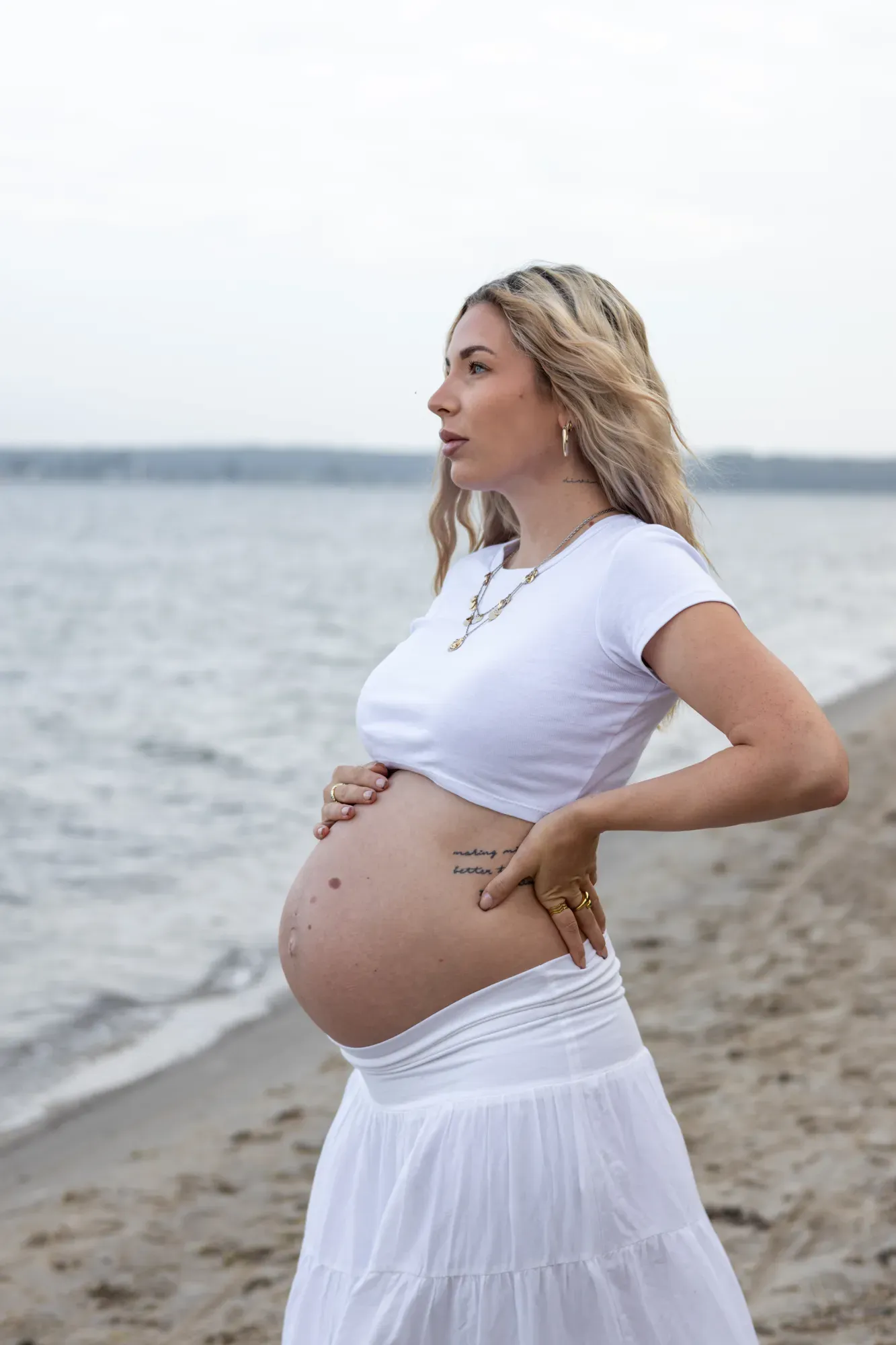 Schwangere Frau am Strand in weißem Rock, die Hände am Babybauch