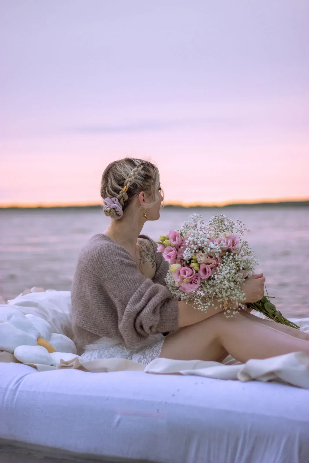 Frau im Strickjäckchen sitzt mit Blumenstrauß auf einem Bett vor rosa Abendhimmel
