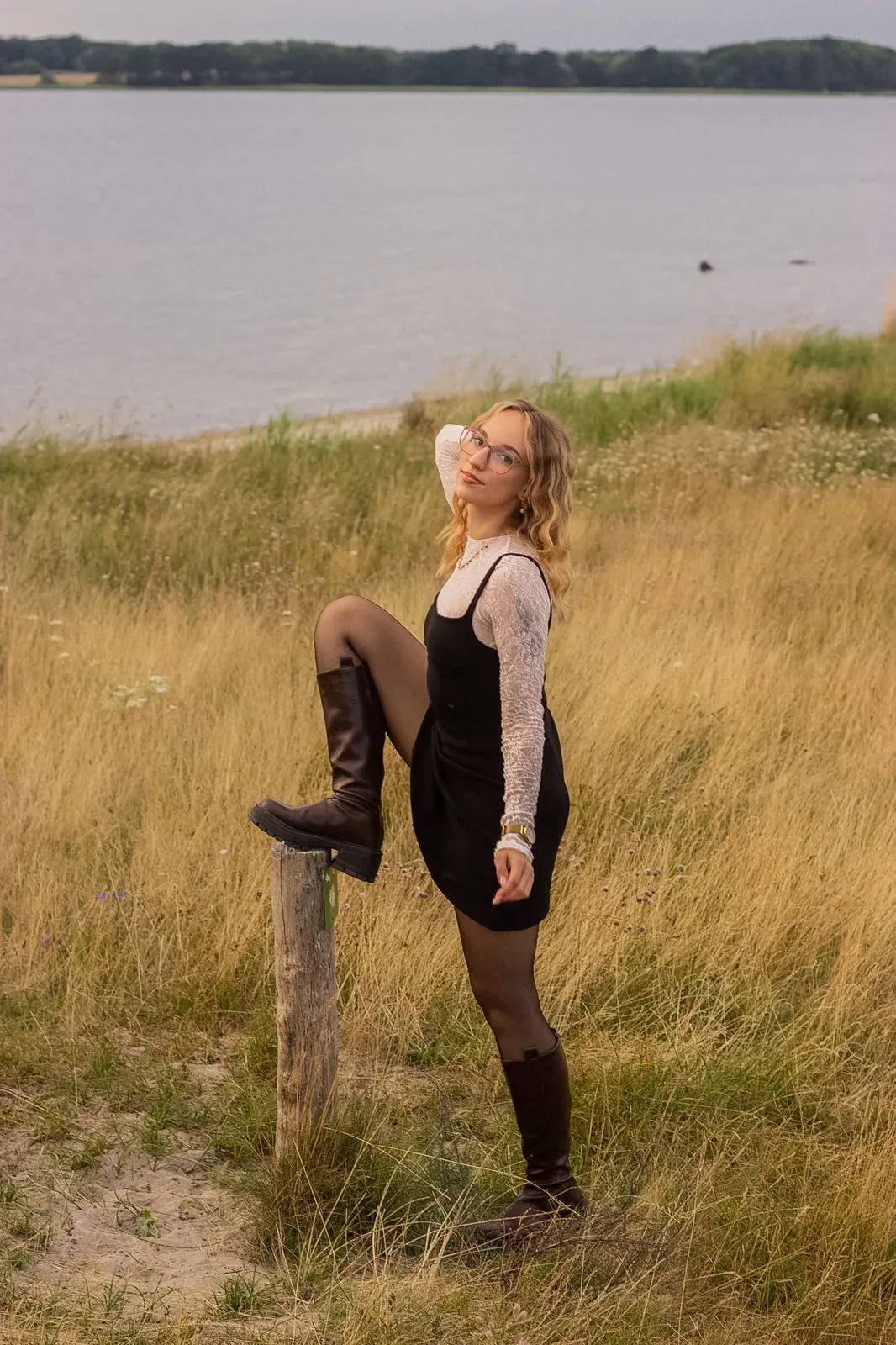 Frau mit Brille steht in hohem Gras am Wasser, ein Bein auf einem Pfahl