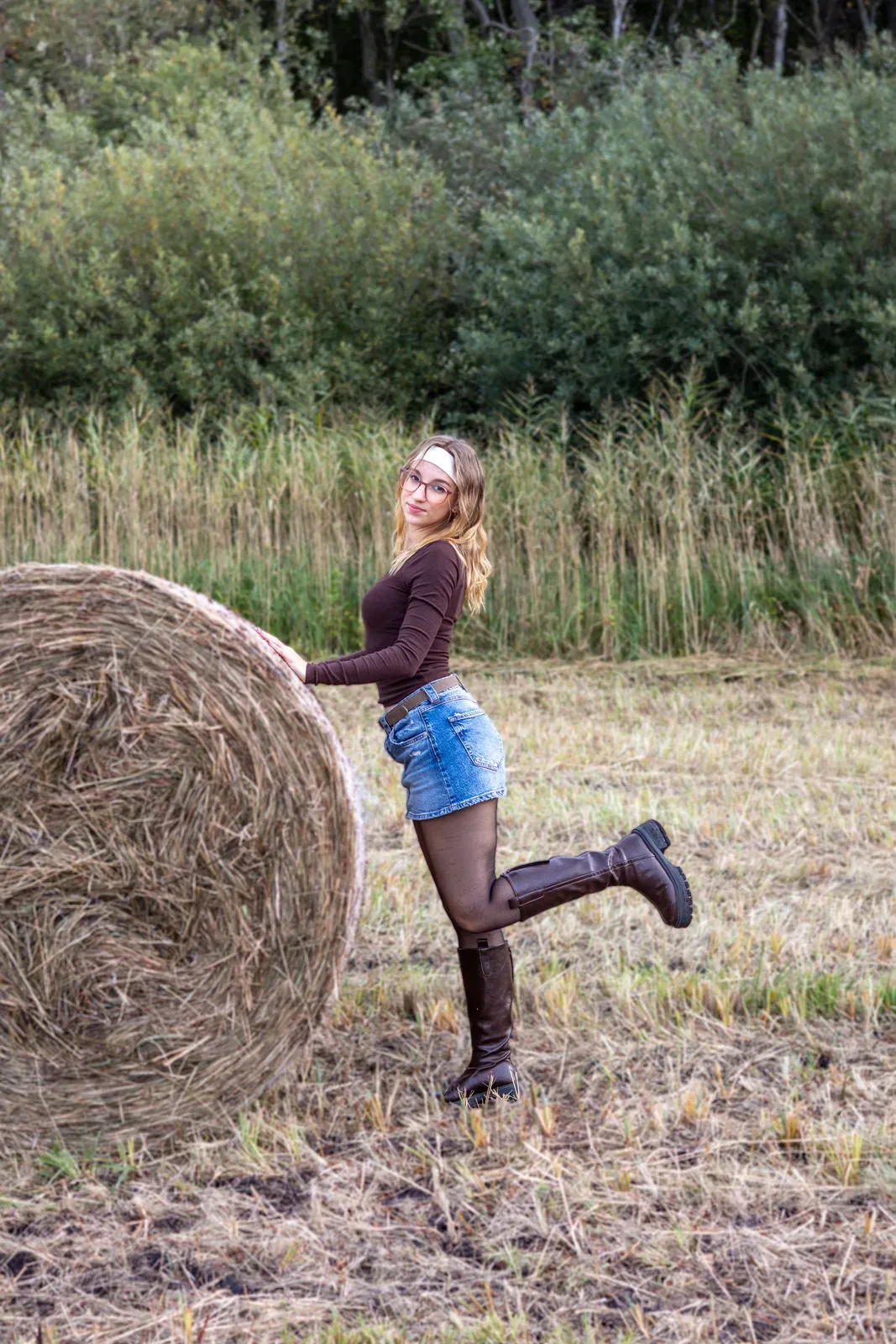 Frau in Jeansrock und Stiefeln lehnt an einem Heuballen auf einem Stoppelfeld