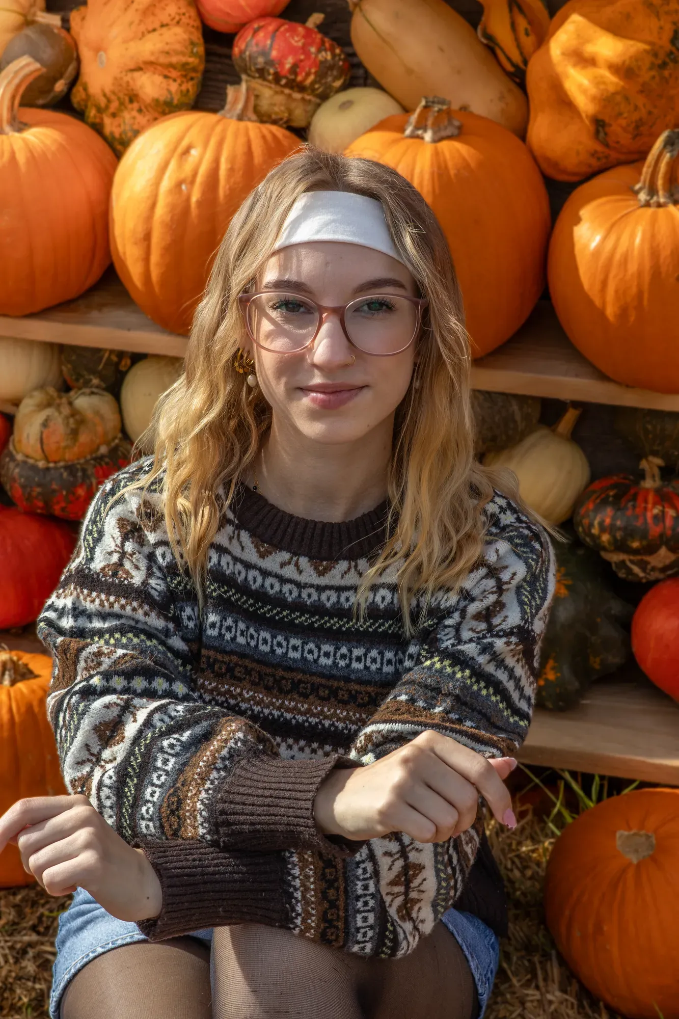 Frau mit Brille und Strickpullover sitzt zwischen Kürbissen und lächelt