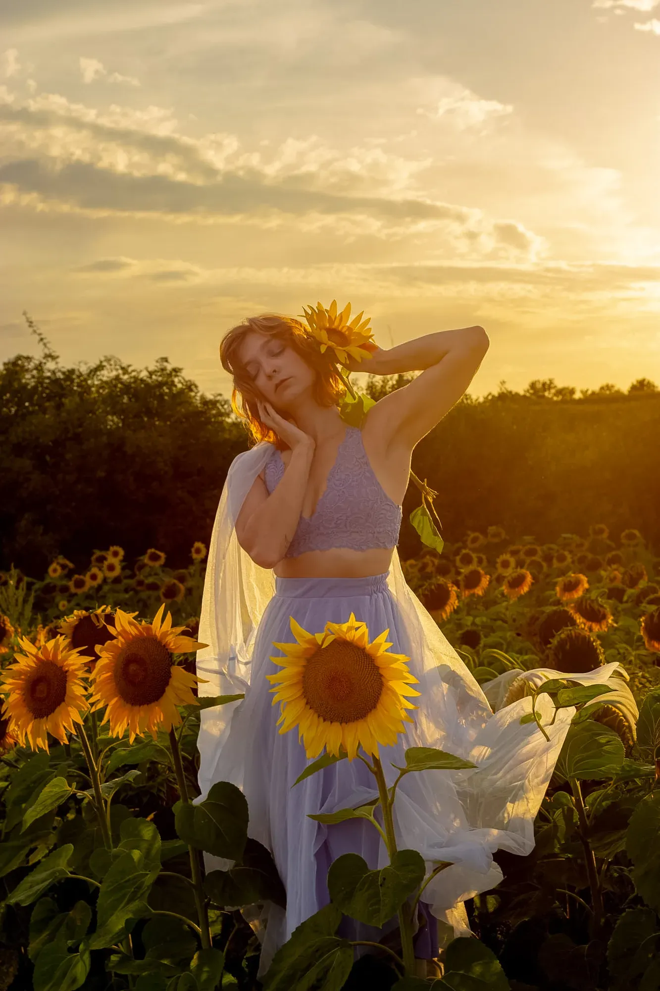 Frau in lilafarbenem Kleid steht im Abendlicht in einem Sonnenblumenfeld